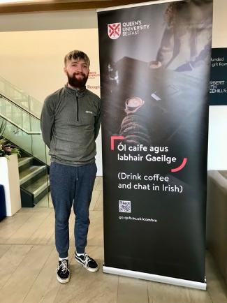 A student stands beside the Comhrá promotional banner that reads: ÓI caife agus labhair Gaeilge - Drink coffee and chat in Irish