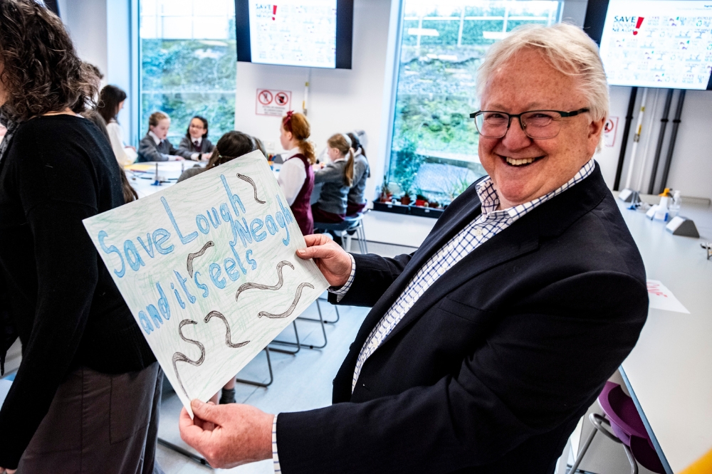 A smiling man in a suit holds a handmade poster that reads 